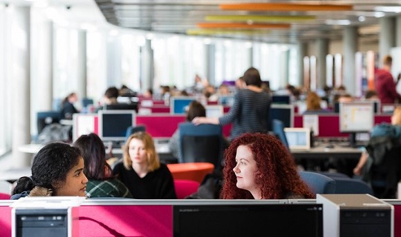 Two QMU students talking whilst working on their desktop computers in a busy classroom