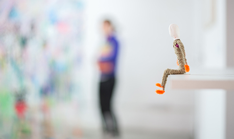 A figure of a person made of pipe cleaners and yarn sitting on the edge of a shelf