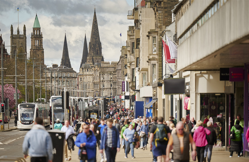 A shot of Edinburgh's Princes Street, featuring some of the shop fronts and passing trams and buses. The street is filled with people.