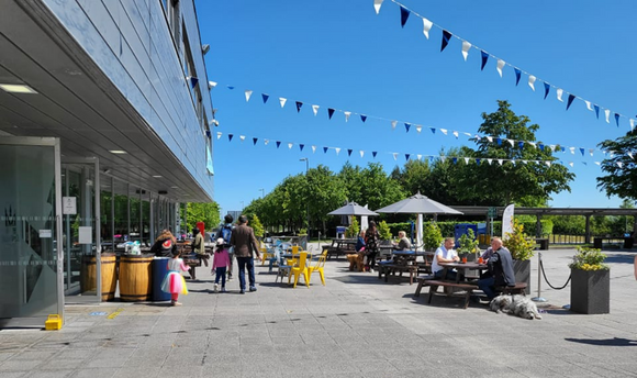 People sitting at picnic tables on a sunny day with blue skies outside Queen Margaret University's Students' Union building