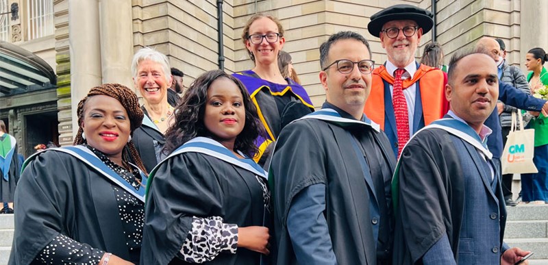 Four students in graduation gowns with family and friends in the background