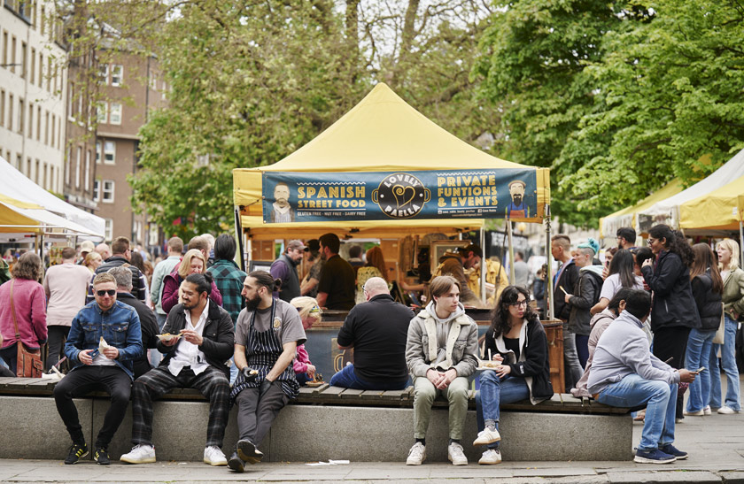 A group of people sitting down on a concrete bench in front of one of the market stalls in Edinburgh's Grassmarket